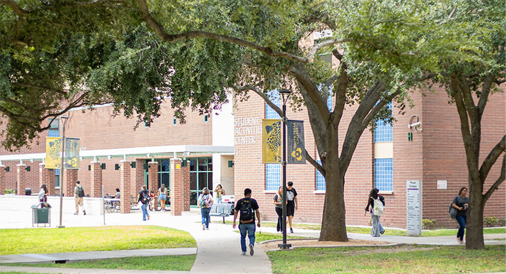 Students walk along shaded concrete pathways surrounding the Student Activities Center at South Texas College, bordered by brick campus buildings and mature trees. STC image